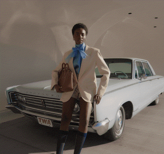 A model standing next to a vintage car wearing shorts, a neutral blazer and bold blue blouse.