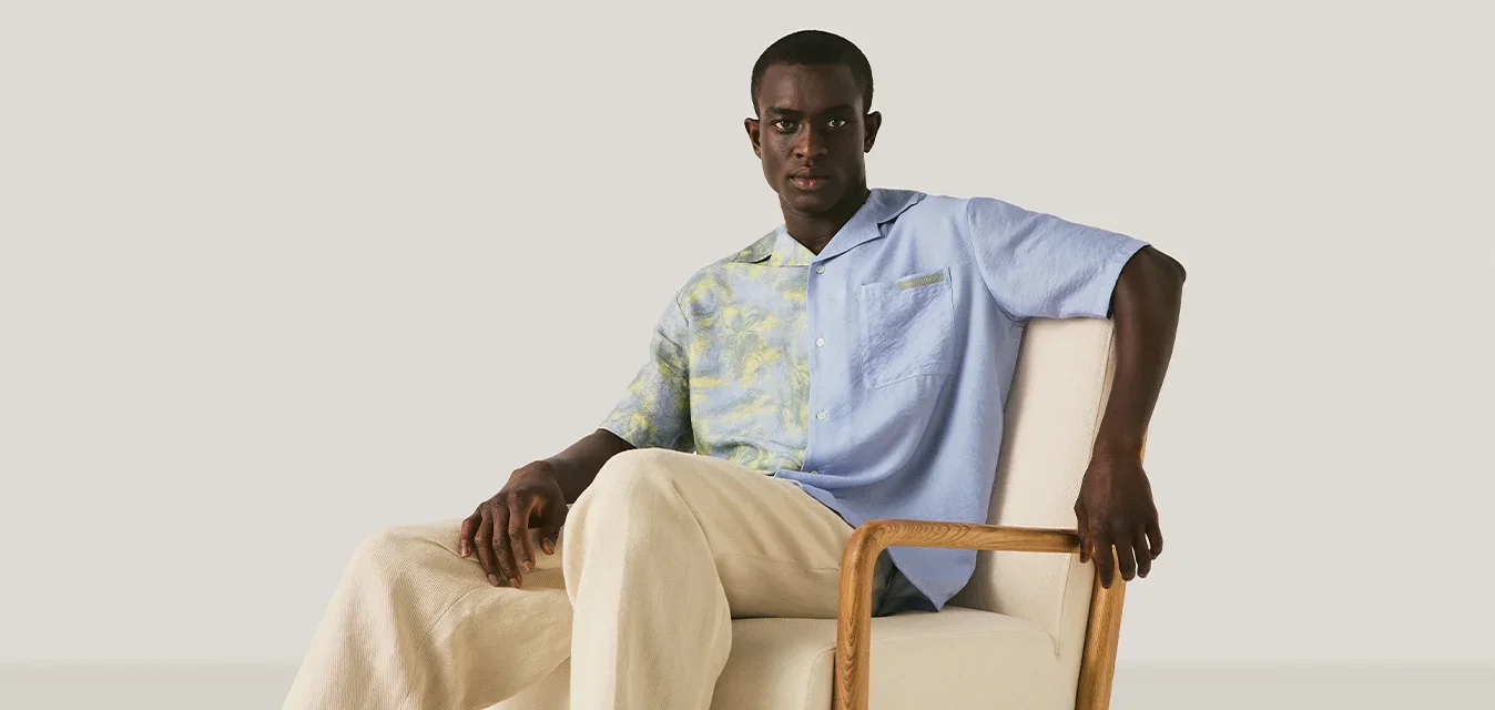 Man in een blauw, patroonhemd en beige broek zittend op een lichtgekleurde gestoffeerde stoel tegen een effen achtergrond.