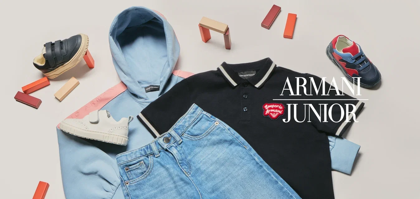 Hellblaue Kapuzenjacke, schwarzes Poloshirt mit rotem Logo, blaue Jeans und drei Paar bunte Kindersneaker rund um die Kleidung auf weißer Oberfläche.
