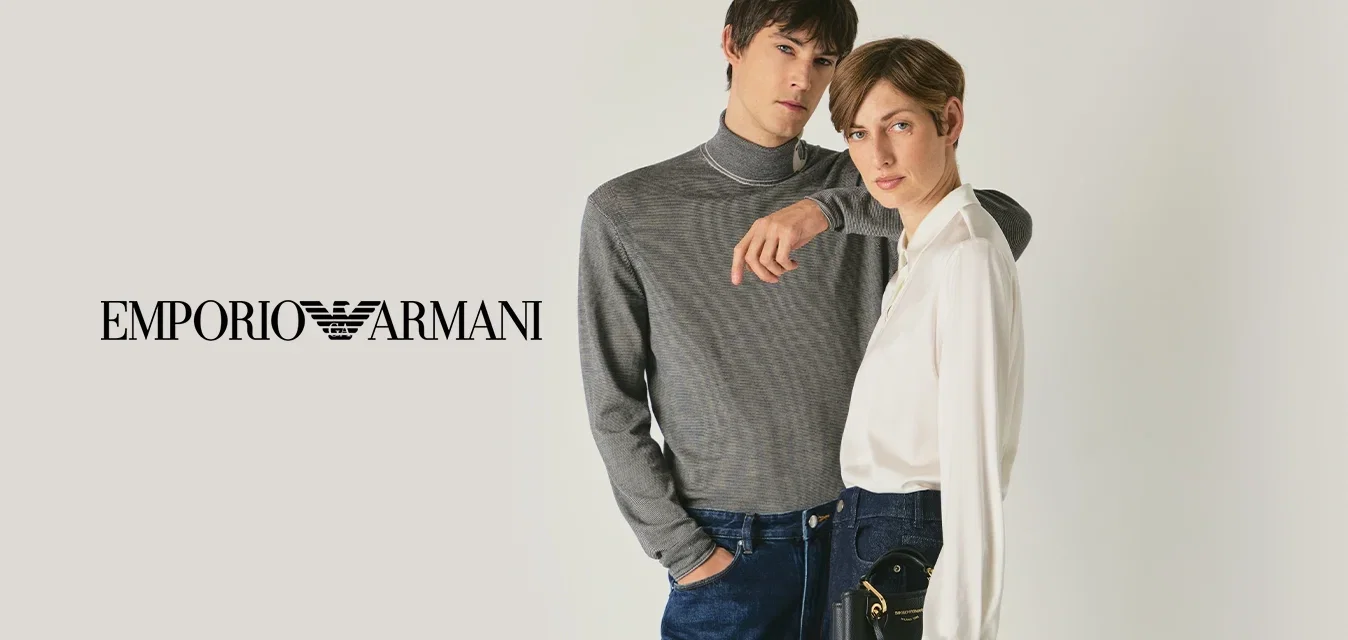 Zwei Models stehen nah beieinander; eines trägt einen grauen gemusterten Rollkragenpullover und Jeans, das andere trägt eine weiße Bluse, weite dunkle Hosen und hält eine schwarze Handtasche.