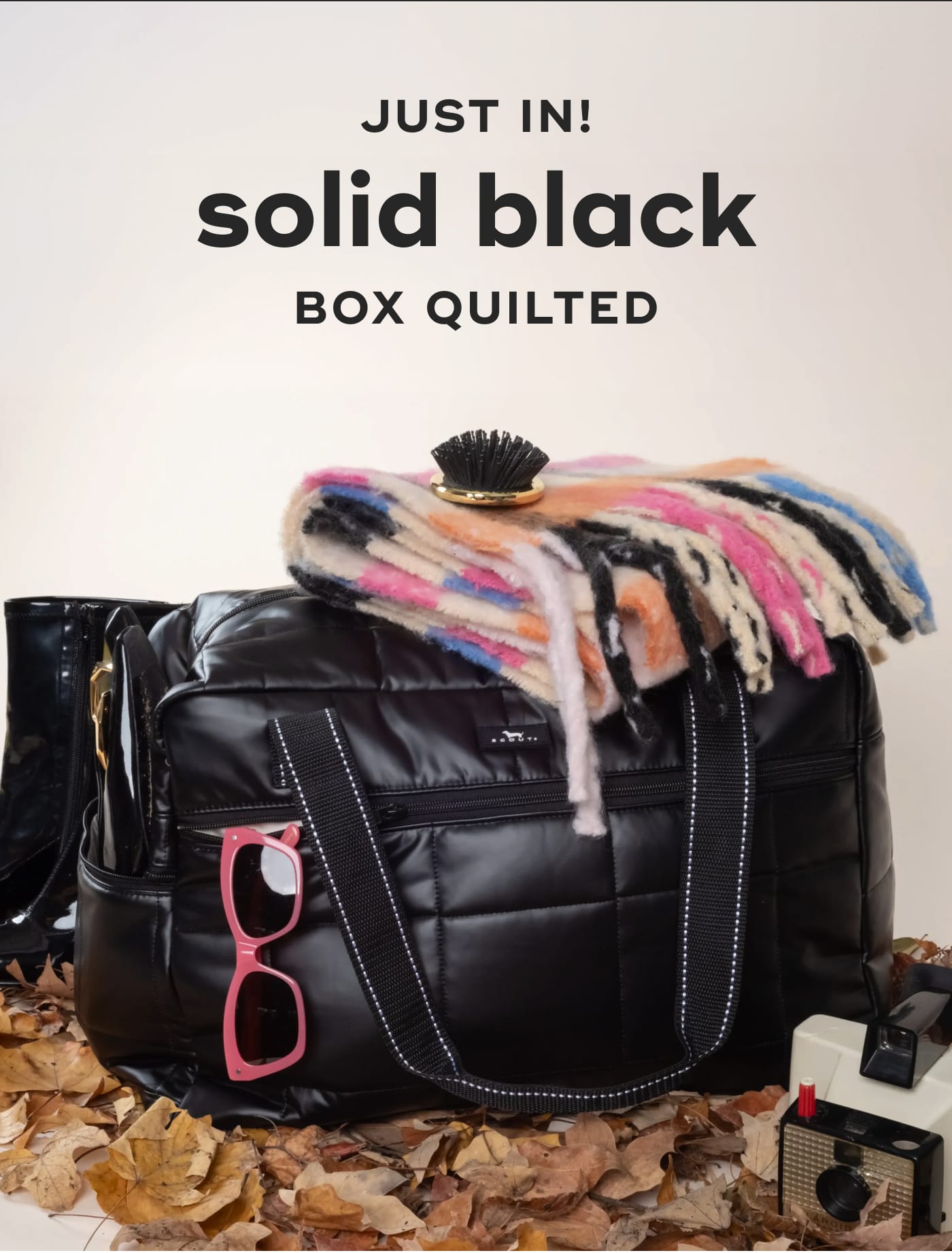 JUST IN! solid black BOX QUILTED. SCOUT bag with a plaid scarf, pink sunglasses, and camera on autumn leaves.