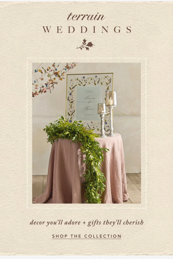 Wedding display table draped in a dusty pink linen tablecloth and lush green garland, holding two candlestick holders and a framed floral welcome sign. Header: terrain WEDDINGS. Subtext: decor you'll adore + gifts they'll cherish. Button: SHOP THE COLLECTION.