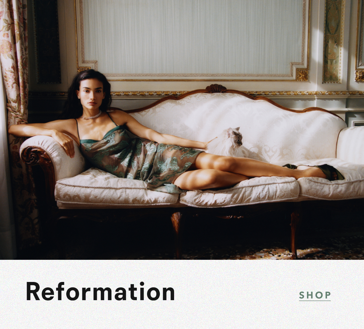 Model reclining on a vintage white velvet sofa next to a fluffy white cat, wearing a green lace-overlay slip dress and heels. Logo: Reformation. Link: SHOP.
