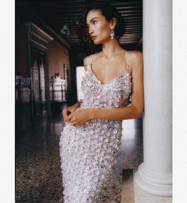 Model leaning against a white marble column in an elegant hallway, wearing an intricately sequined and bead-embellished ivory gown with spaghetti straps and drop earrings.