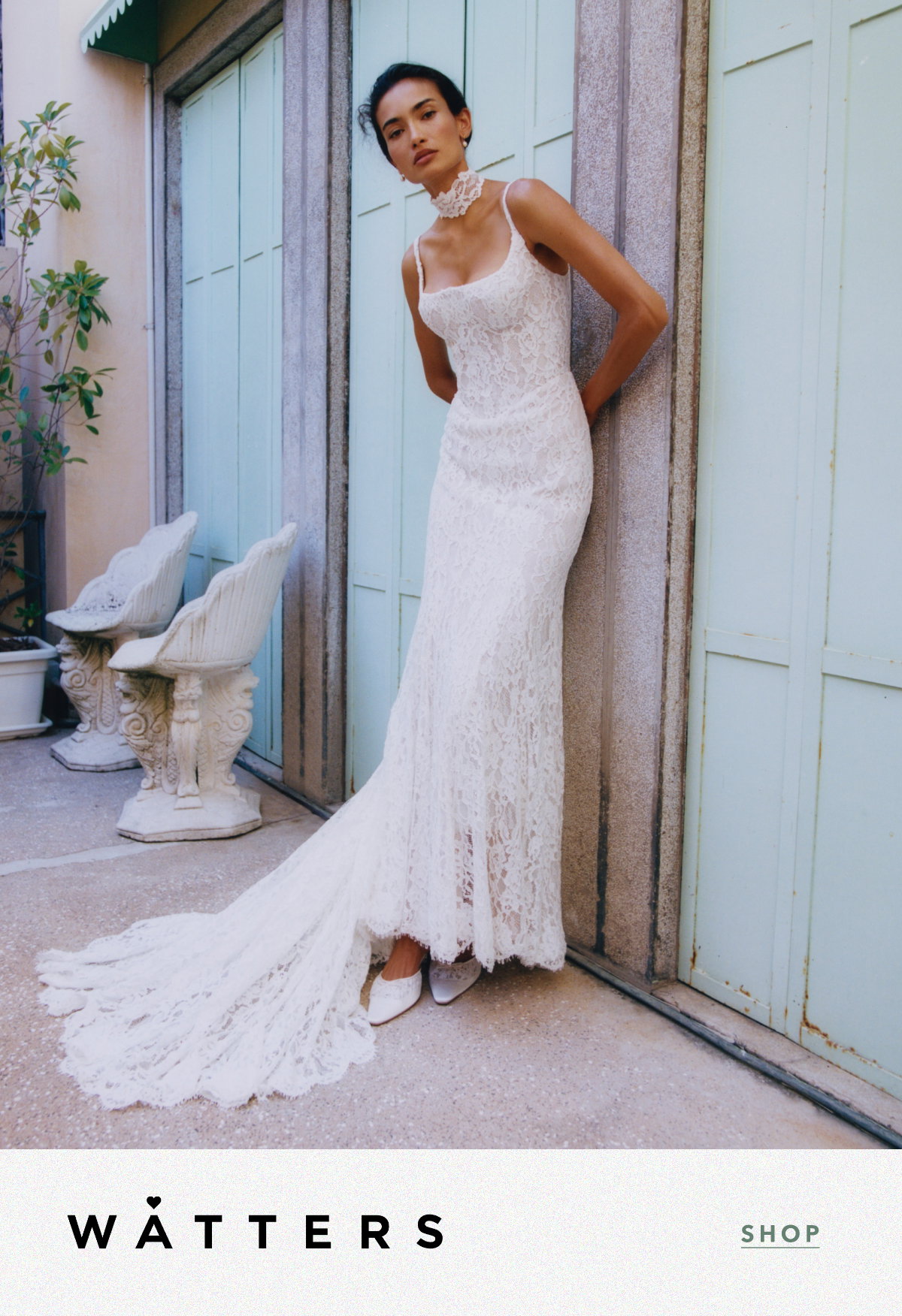 Model wearing a floor-length white lace bridal gown with a sweeping train, spaghetti straps, and a matching lace choker, leaning against a stone doorway. Logo: WATTERS. Link: SHOP.