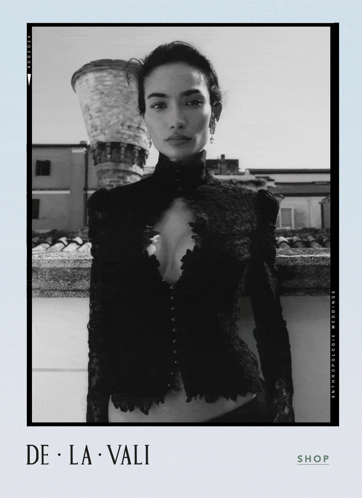 Black-and-white framed portrait of a model on a rooftop wearing a high-neck, cut-out black lace long-sleeve top. Logo: DE · LA · VALI. Link: SHOP.