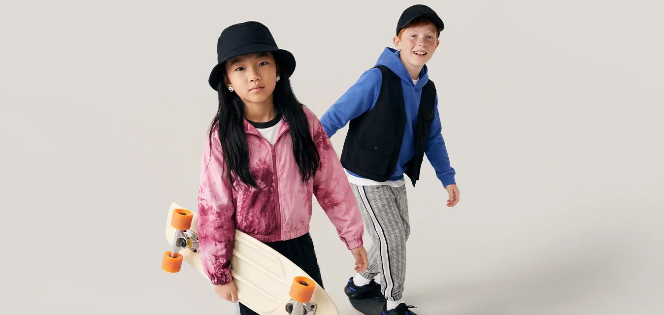 Twee kinderen met skateboards, een staat en houdt een skateboard vast, de ander rijdt op een skateboard, beiden dragen casual kleding en hoeden.