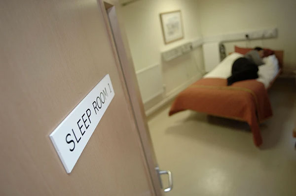 A patient sleeps in a bed, viewed through an open doorway at a sleep clinic.