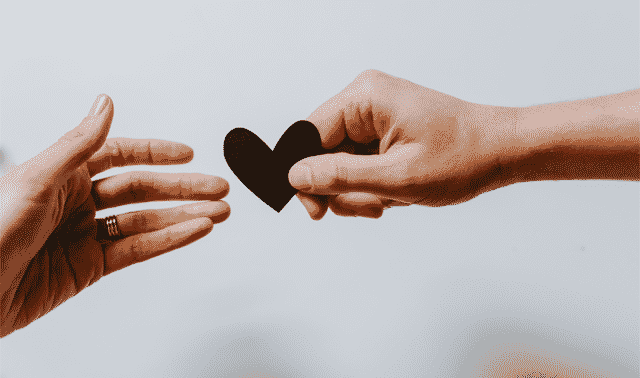 Two hands reach towards each other, with one hand holding a small black heart cutout.