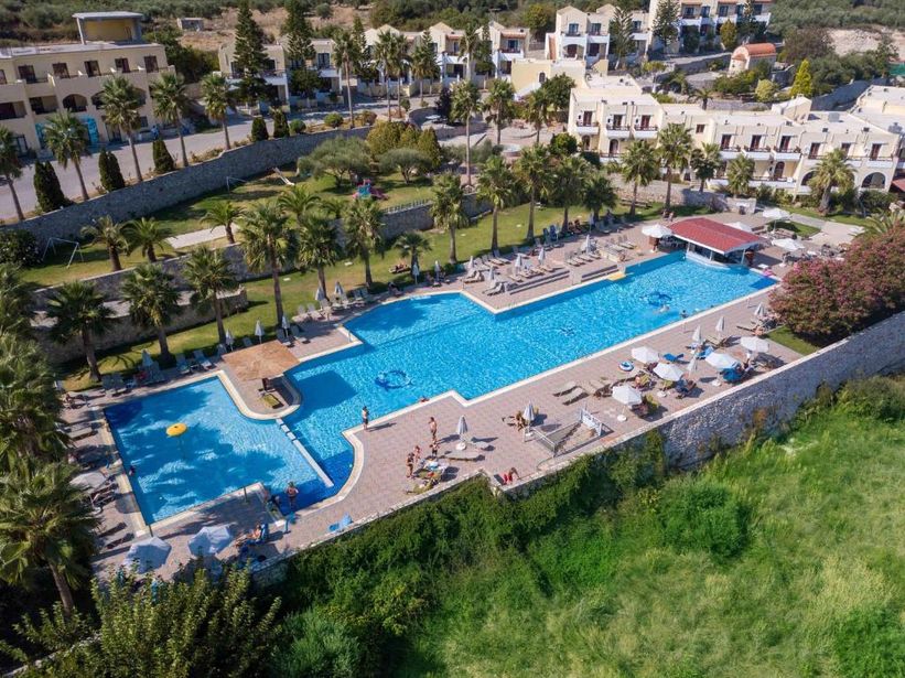 Aerial view of a sunlit resort swimming pool with sunloungers, umbrellas and palm trees beside Mediterranean-style hotel buildings