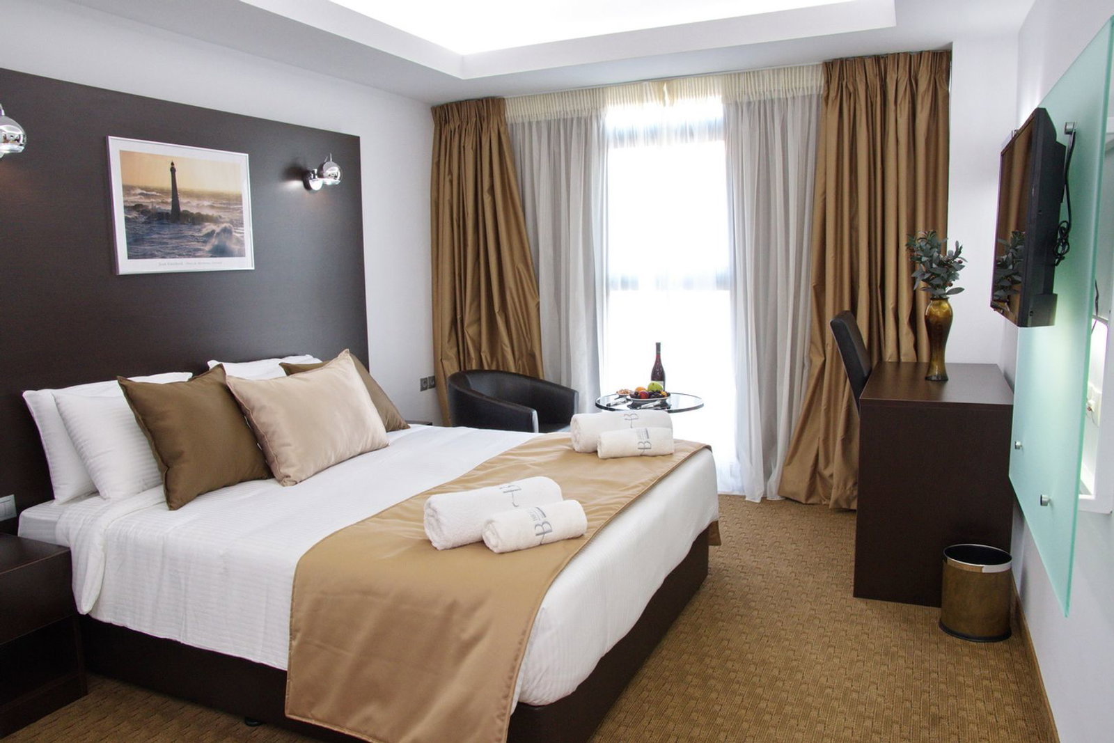 Modern hotel room with double bed, beige bed runner and pillows, rolled towels on bed, window with curtains, armchair, desk and wall-mounted TV
