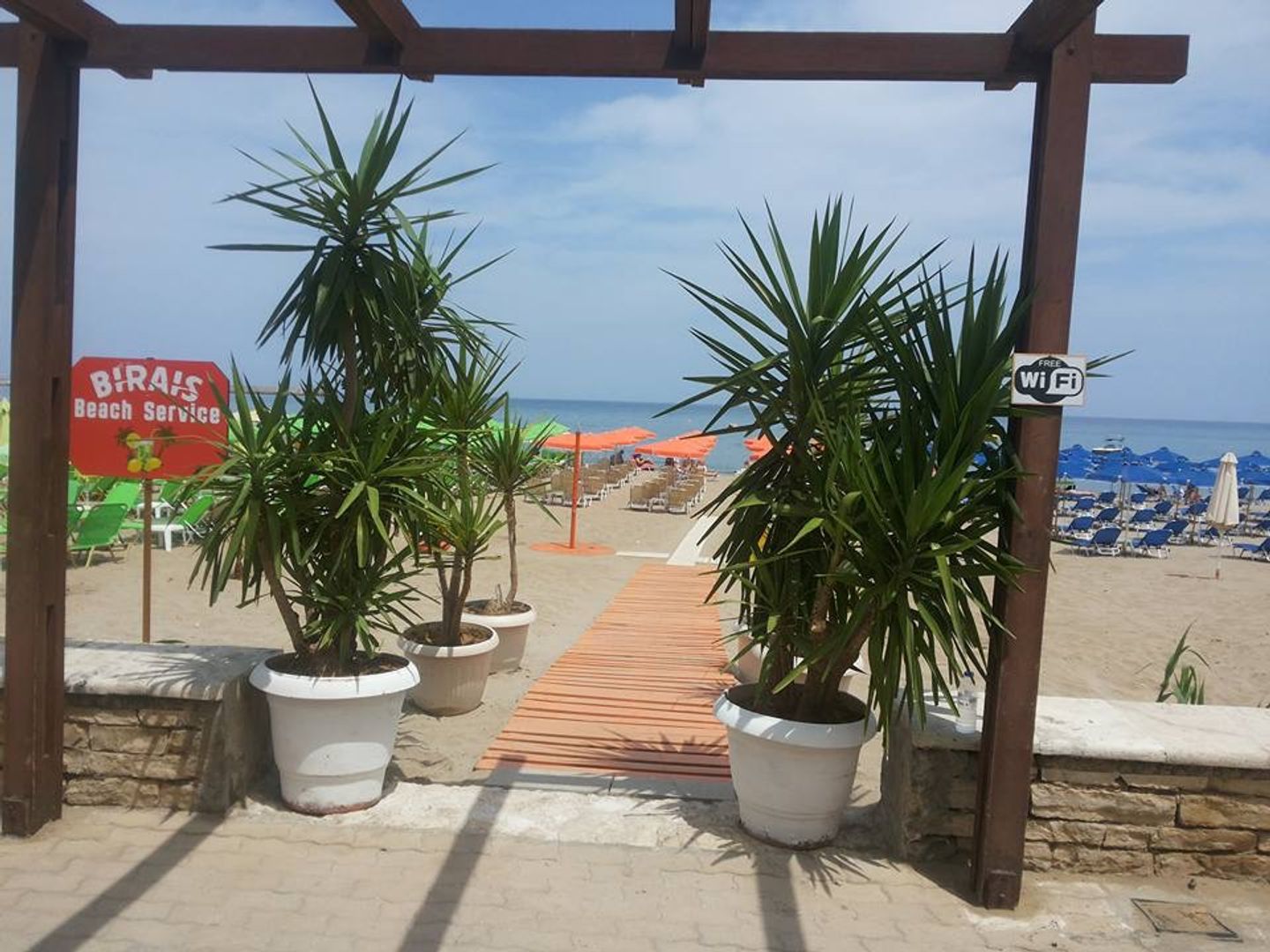 Wooden pergola entrance with potted palm plants and boardwalk leading to sandy beach with sunbeds and colourful umbrellas