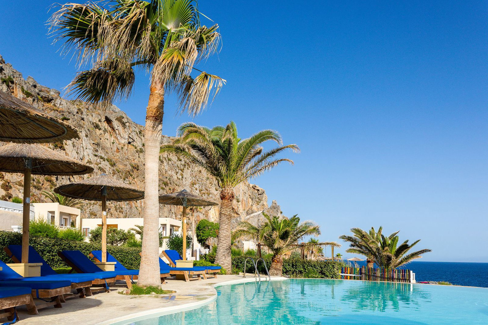 Infinity pool at a coastal resort with palm trees, blue sun loungers and thatched parasols overlooking sea and rocky cliffs.