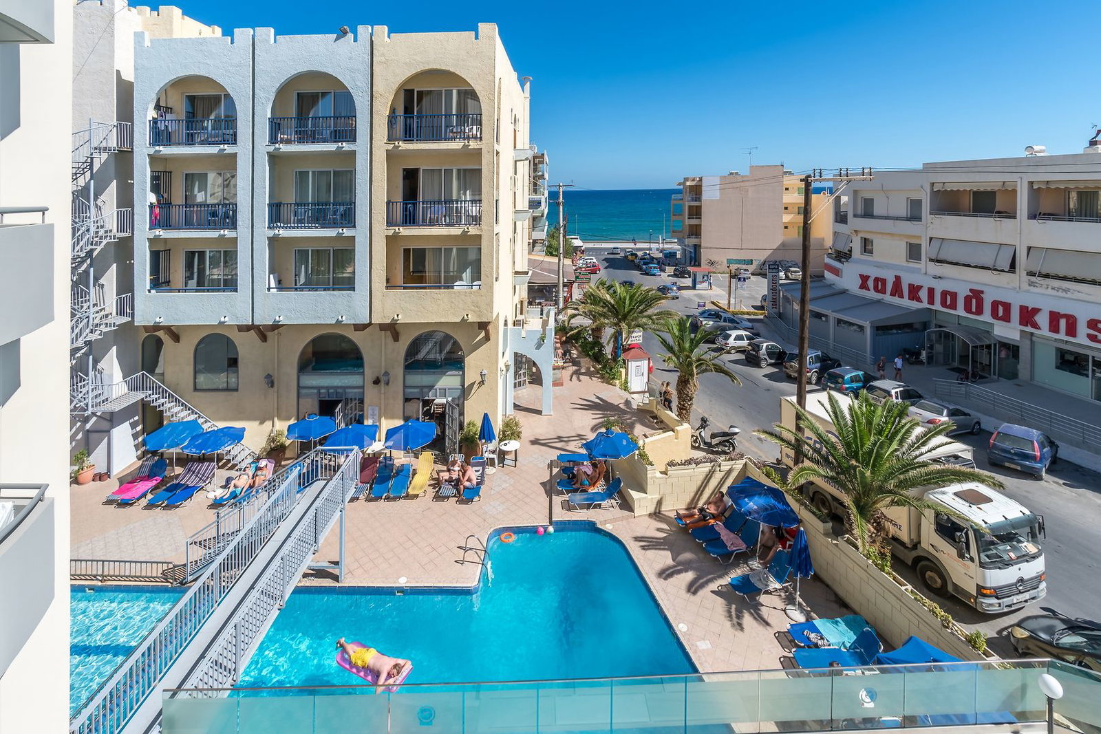 Seaside hotel courtyard with outdoor pool, sunbeds and blue umbrellas, guest on a pink float, palm trees and sea view beyond