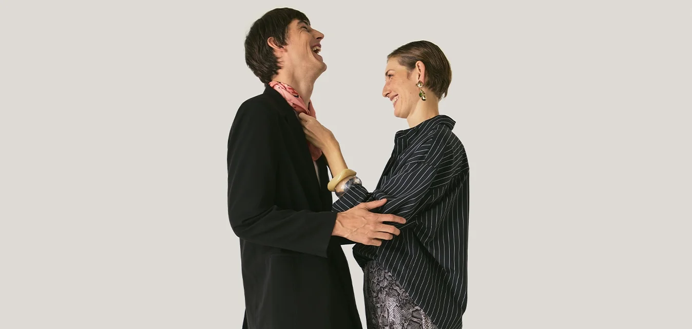 Twee mensen lachen en staan tegenover elkaar, terwijl de een de sjaal van de ander ajusteert. Beiden dragen kleding met patronen en donkere kleuren, op een effen lichte achtergrond.