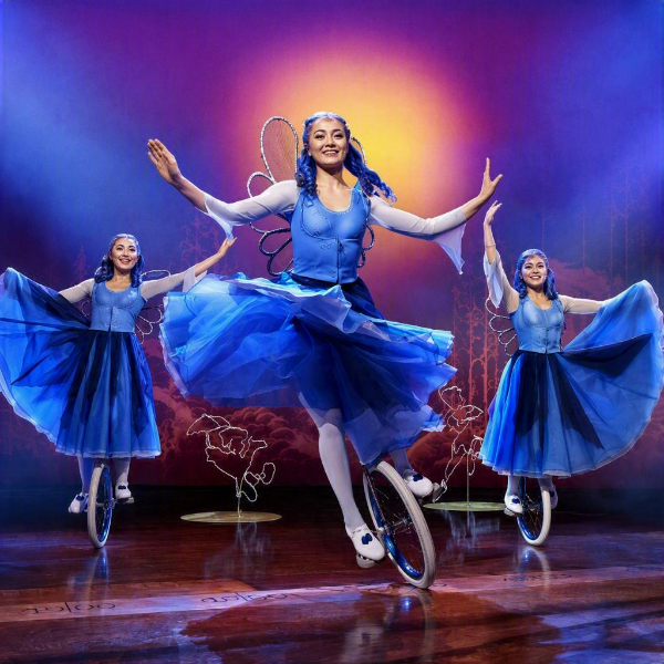 A young woman in a bright blue fairy-like costume rides a unicycle on a theater stage, with two mirrored versions of herself performing alongside. The performers wear flowing blue tulle skirts, white tights, and decorative wings, creating a whimsical fantasy look. Warm stage lighting and a glowing circular backdrop contrast with the cool blue costumes, enhancing the magical atmosphere. The polished wooden floor and stylized background props suggest a professional stage show or circus-style performance.
