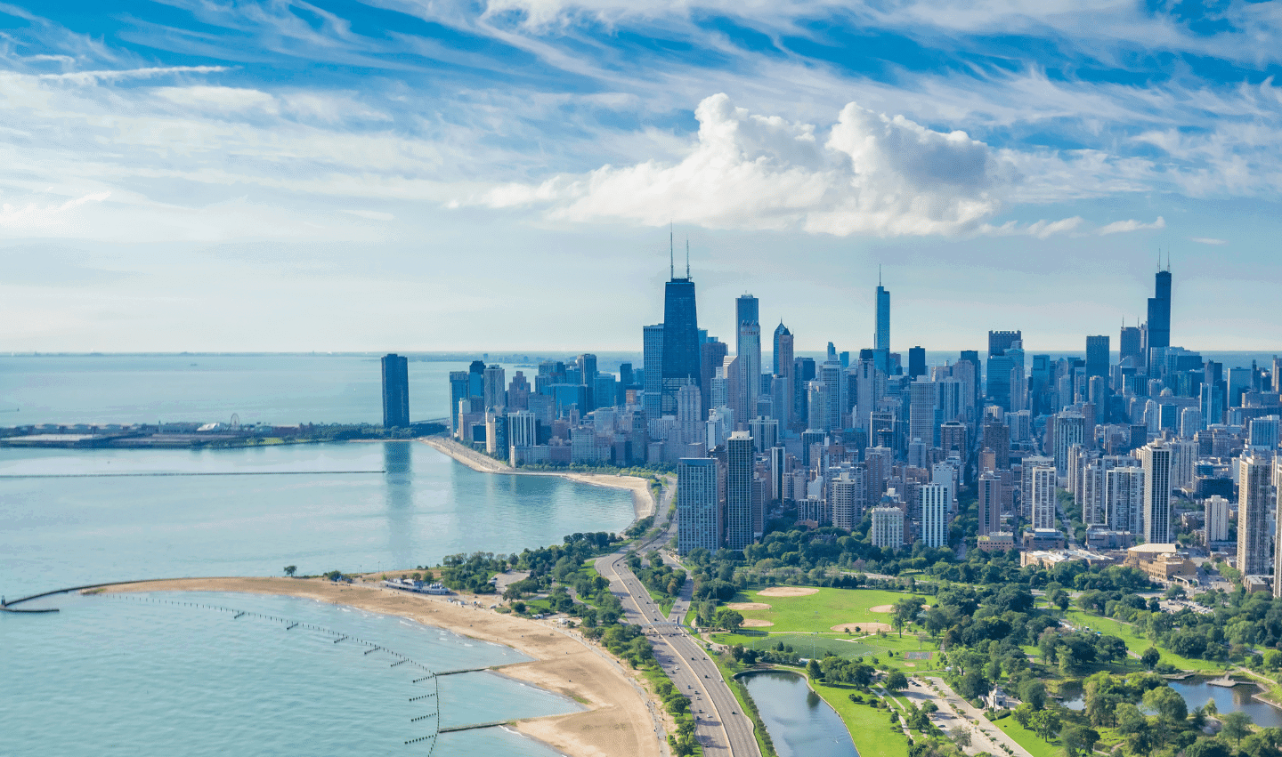 Aerial view of Chicago skyline with tall buildings, a large lake, sandy beaches, green parks, and a curving road along the shoreline under a partly cloudy sky.