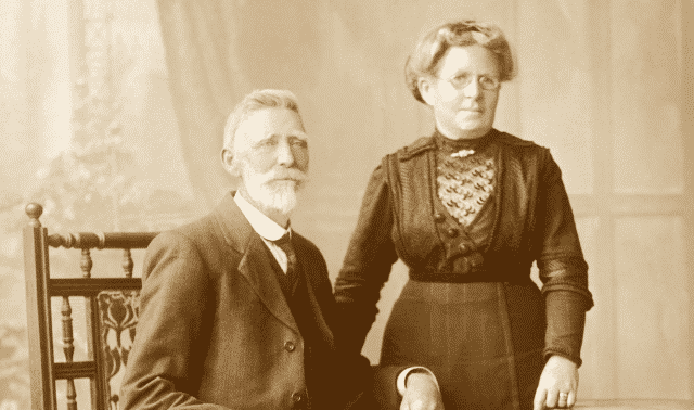 A vintage sepia-toned portrait of an older man with a beard seated beside an older woman in a dark dress standing, both looking at the camera.