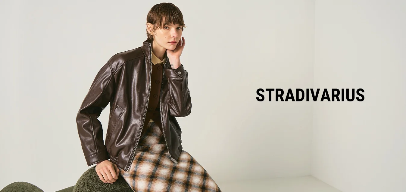 Person trägt eine dunkelbraune Lederjacke und einen karierten Rock, sitzt auf einem grünen, strukturierten Stuhl vor einem schlichten hellen Hintergrund.