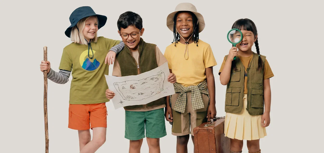 Vier Kinder, verkleidet als Entdecker, halten eine Karte, einen Wanderstock, einen Koffer und eine Lupe, lächeln und stehen zusammen.