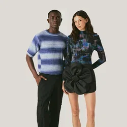Ein Mann in einem blau gestreiften Pullover und schwarzen Hosen steht neben einer Frau, die ein langärmeliges gemustertes Oberteil und einen schwarzen Rock mit einem großen Blumendetail trägt.