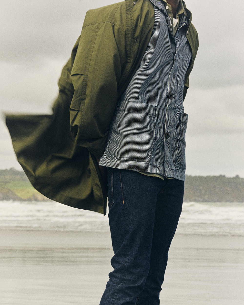 Image of woman on beach with sand covered hands. She wears Rinse Wash denim jeans, green shirt, and green sleeveless jumper.