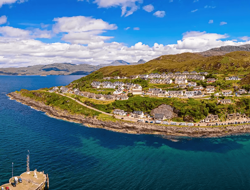 A scenic coastal view featuring a village along a green hillside, with blue waters and mountains in the background.