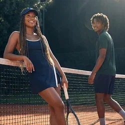 Zwei junge Personen stehen auf einem Sand-Tennisplatz am Netz, eine lehnt sich mit einem Schläger daran, im Hintergrund Sonnenlicht und Bäume.