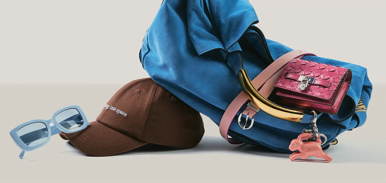 Blaue Handtasche mit einem pinkfarbenen Portemonnaie und Anhänger am Griff, neben einer braunen Baseballkappe und blauen Sonnenbrillen auf einer flachen Oberfläche.