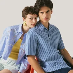 Zwei Personen sitzen dicht nebeneinander auf einem roten Stuhl, eine trägt ein blau gestreiftes Hemd und dunkle Shorts, die andere ein hellblaues Hemd mit weißen Shorts.
