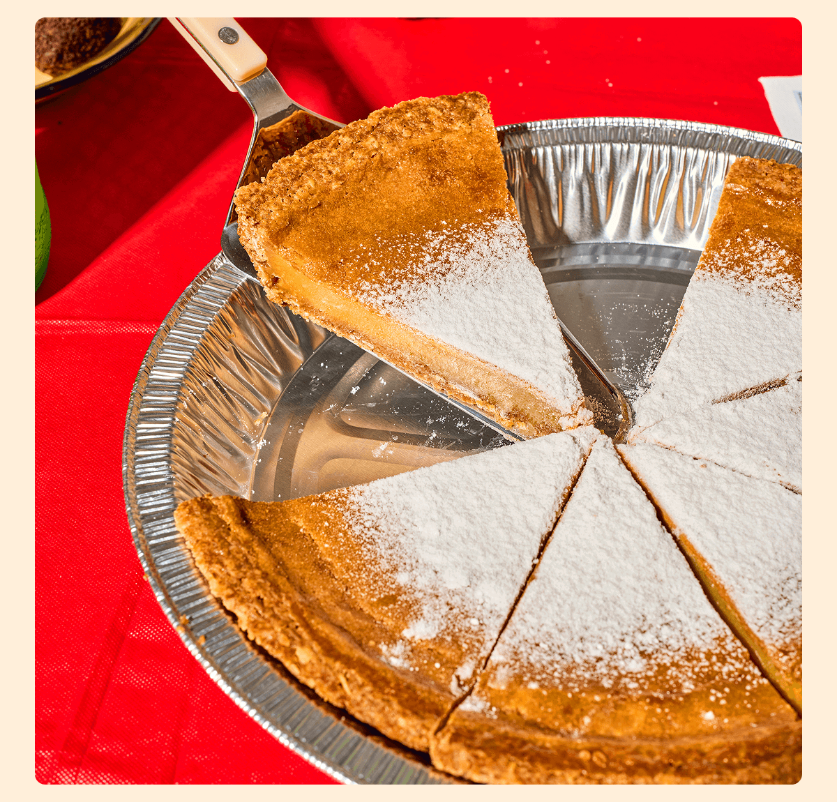 A slice of golden Milk Bar Pie dusted with powdered sugar is lifted from a partially sliced pie in a foil tin on a bright red tablecloth.