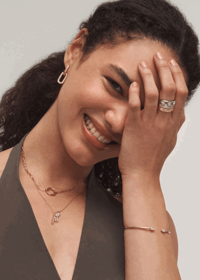 Woman smiling while wearing layered gold necklaces, gold drop earrings, rings and a gold bangle bracelet.