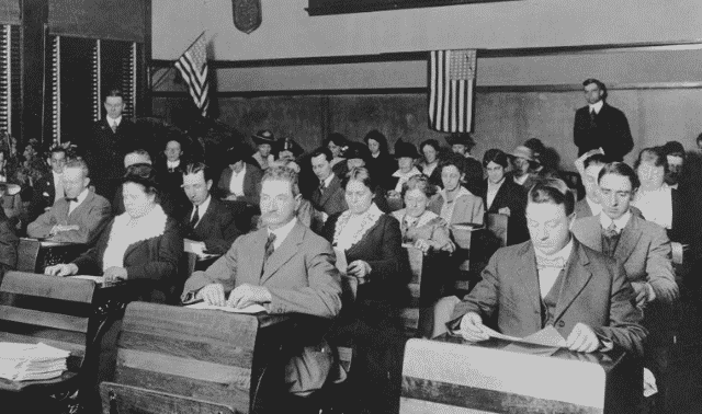 A group of men and women seated in a classroom-like setting, some reading papers. American flags are displayed on the walls in the background.