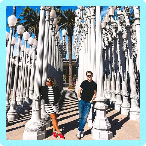 Image description: Two people standing in Chris Burden’s Urban Light installation at LACMA