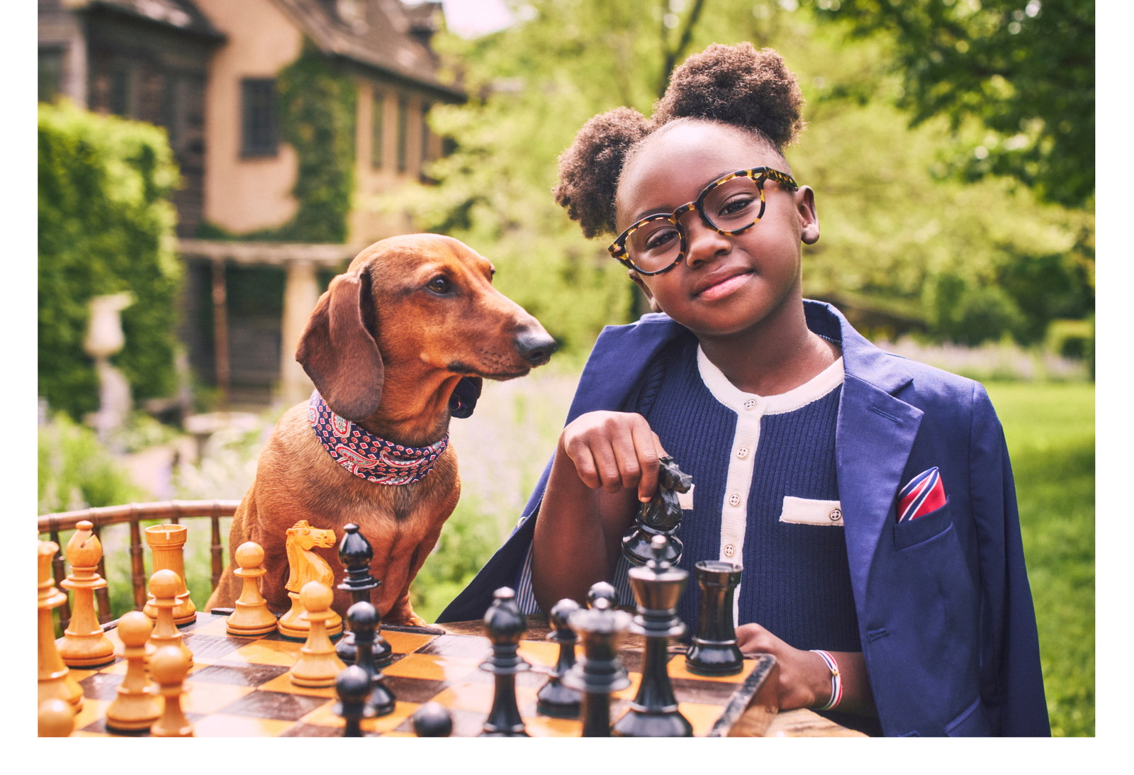 A young girl wearing glasses and a navy blazer plays chess outdoors with a dachshund wearing a paisley bandana