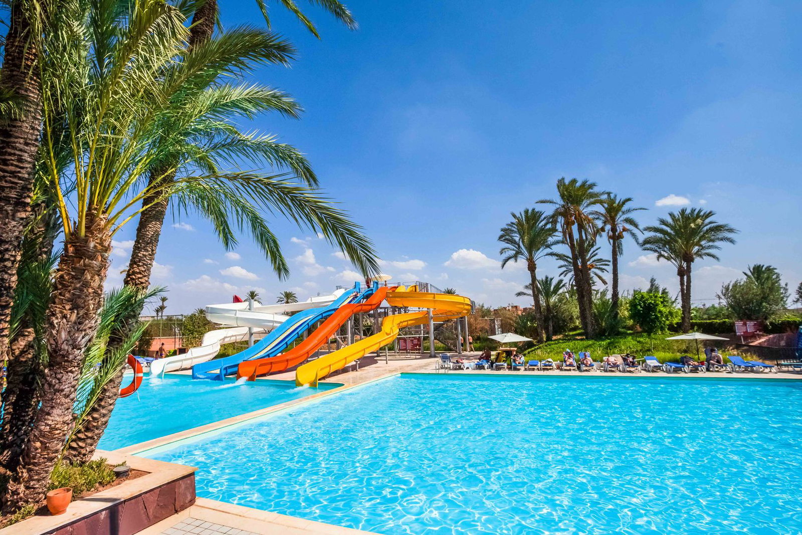 Outdoor resort swimming pool with bright blue water, colourful water slides, palm trees and sun loungers under a clear sky