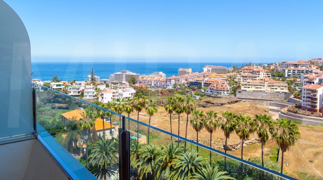 Sea-view balcony with glass railing overlooking palm trees, terracotta-roofed coastal town and blue ocean