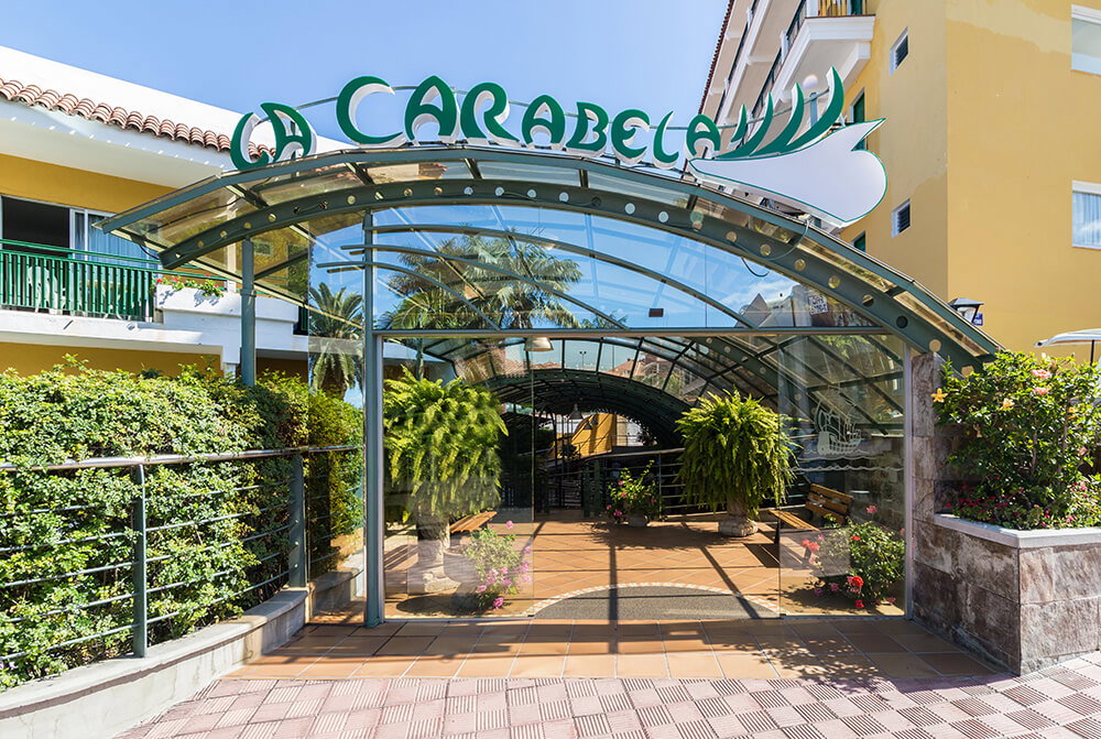 Glass‑canopied entrance to La Carabela hotel with potted palms, tiled walkway and tropical plants under a clear blue sky
