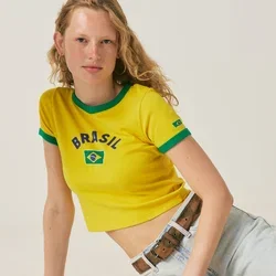Junge Frau in einem gelben Brasilien-T-Shirt und Jeansshorts sitzt auf dem Boden neben einem schwarzen Standventilator in einem schlichten weißen Raum.