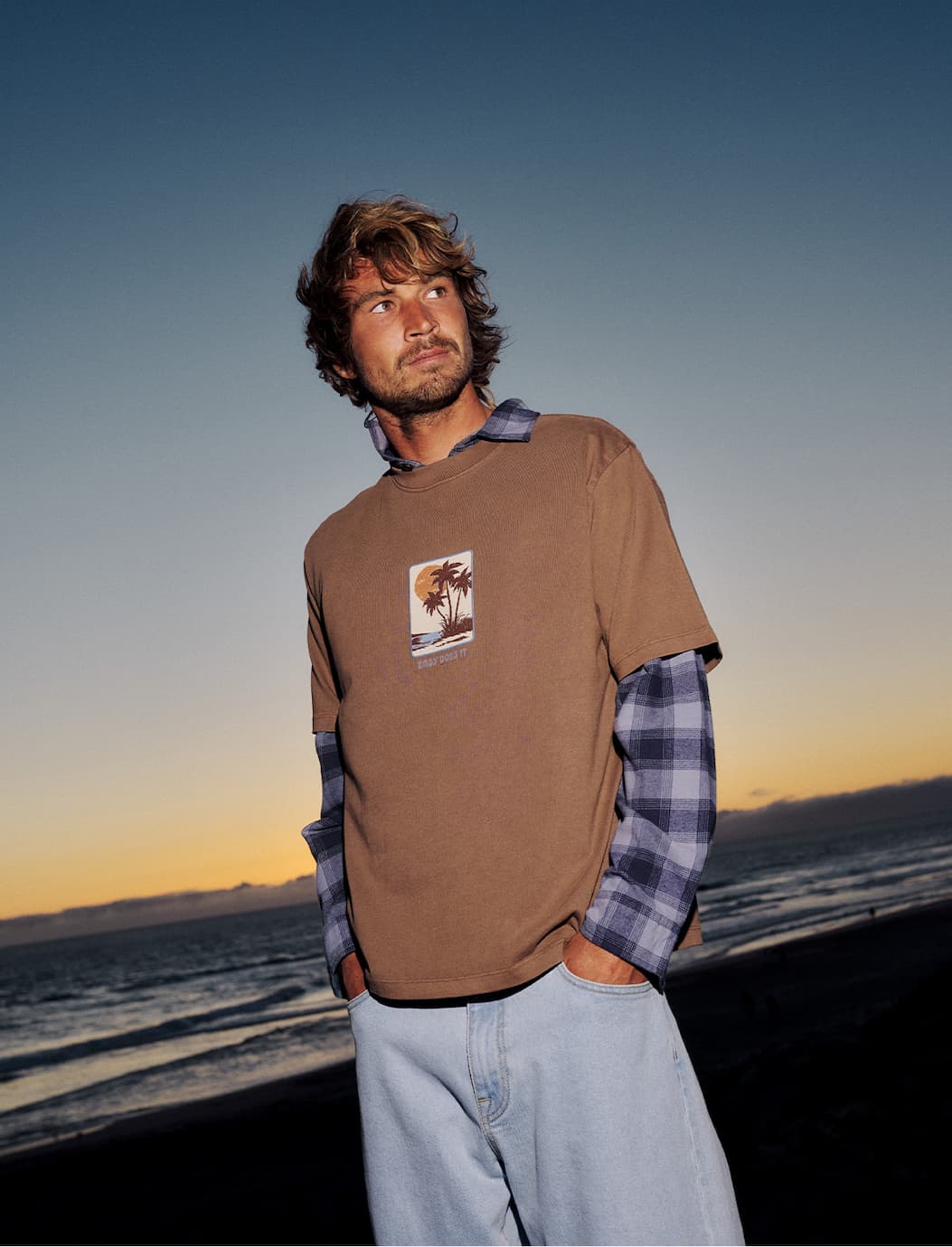 Male model wearing the Brisa Box Fit Tee standing on the beach at sunset.