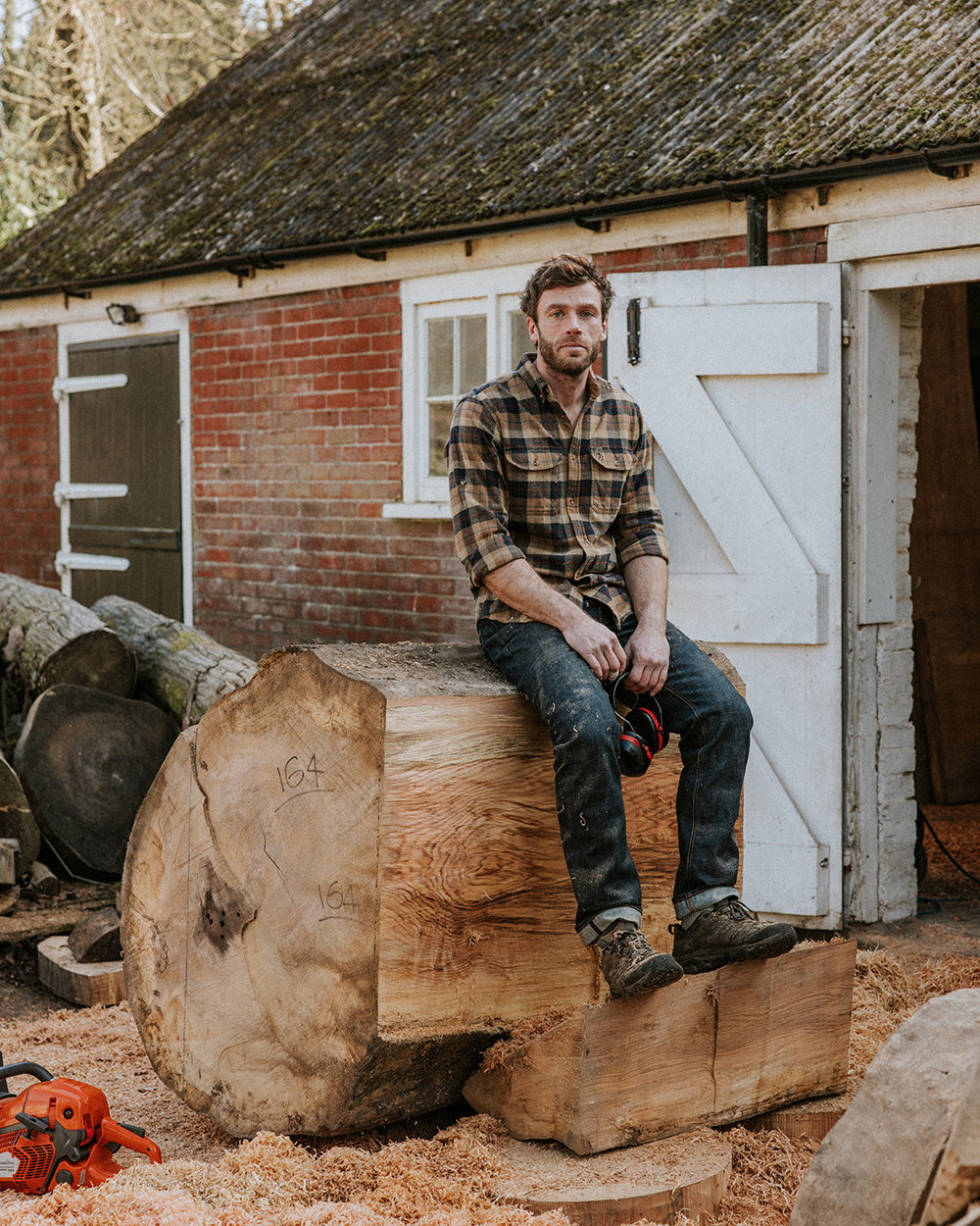 Oliver Chalk sits on a log of wood. In plaid shirt, Hiut jeans, and work boots. Outside of his workshop.