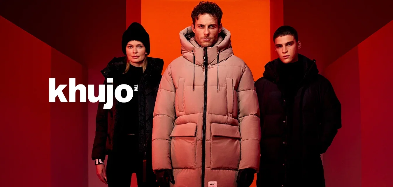 Drie mensen in winterjassen en laarzen staan in een rood verlichte gang met geometrische muren en vloer.