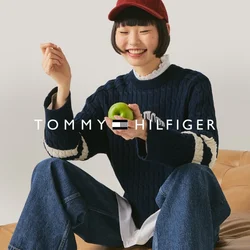 Junge Person mit roter Mütze, marineblauem Pullover und Jeans sitzt auf einem hellbraunen Ledersofa, hält einen grünen Apfel und lächelt vor einem schlichten Hintergrund.
