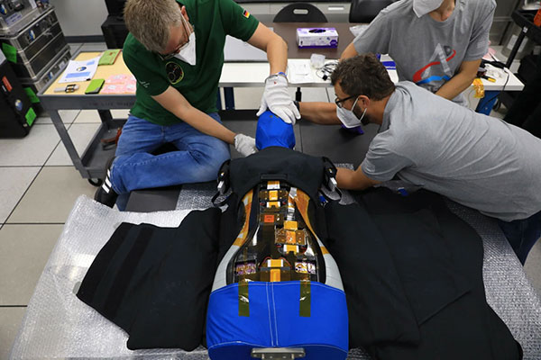 Three technicians assemble a vest on a human-shaped, blue-and-black robotic device with exposed electronics in a laboratory setting.