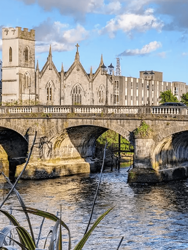 A stone bridge arches over a river, with a historic church and modern buildings in the background.