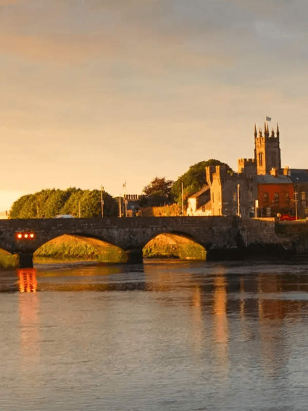 A stone bridge over a river at sunset, with buildings and trees in the background.