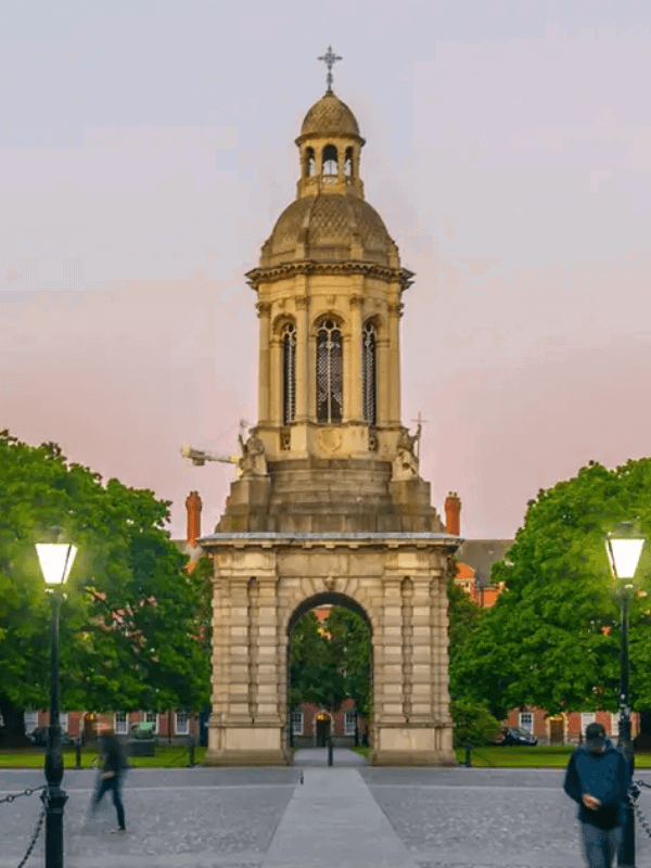 Historic bell tower framed by lush green trees at dusk, with people walking nearby.