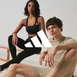 Twee jonge volwassenen in sportkleding zitten bij houten banken in een minimalistische binnenruimte, beide dragen Adidas-sneakers.