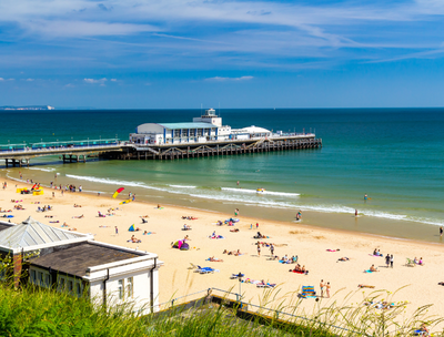 A sunny beach scene with people relaxing on the sand and a pier extending into the sea.
