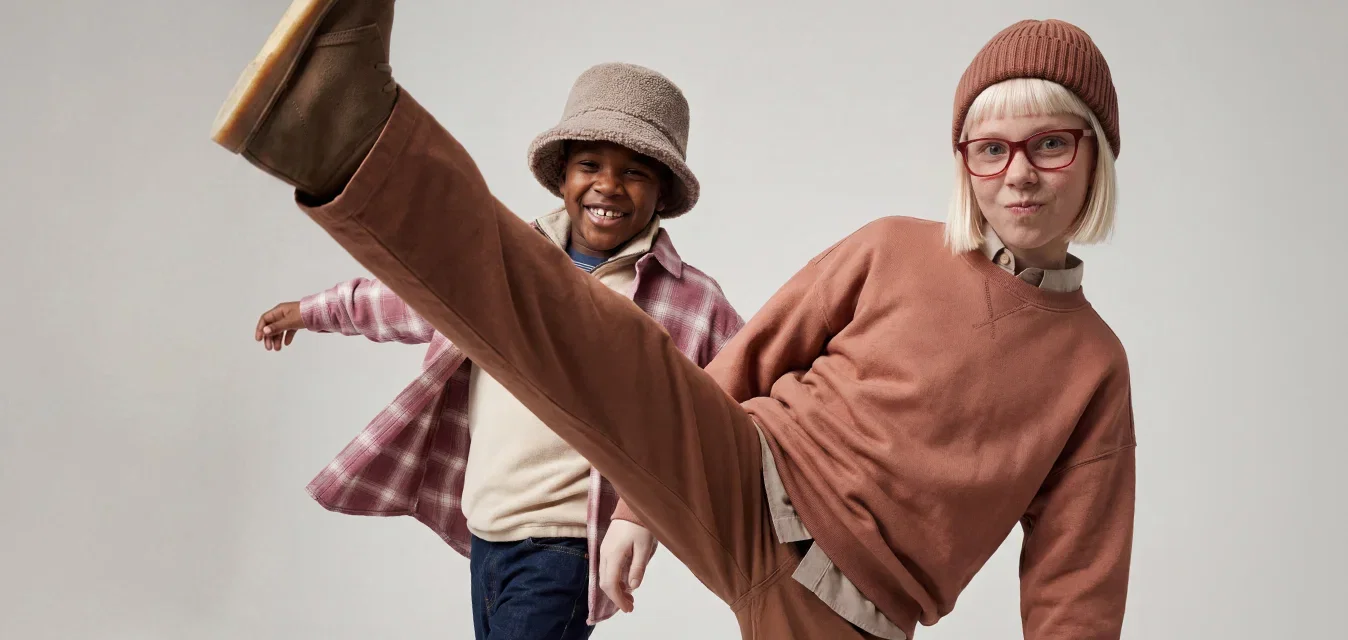Zwei Kinder tanzen in Freizeitkleidung, eines kickt hoch, trägt ein braunes Outfit und eine Mütze, das andere lächelt in einem karierten Hemd und Hut.